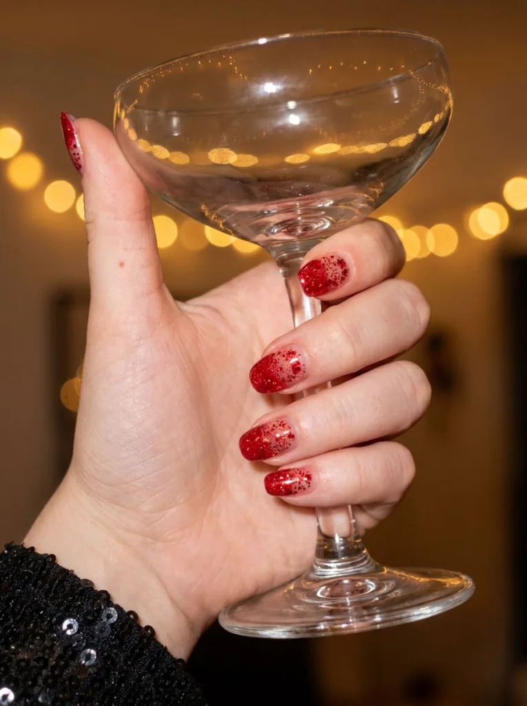 Hand with red glitter nails holding the stem of a champagne glass at a holiday party.