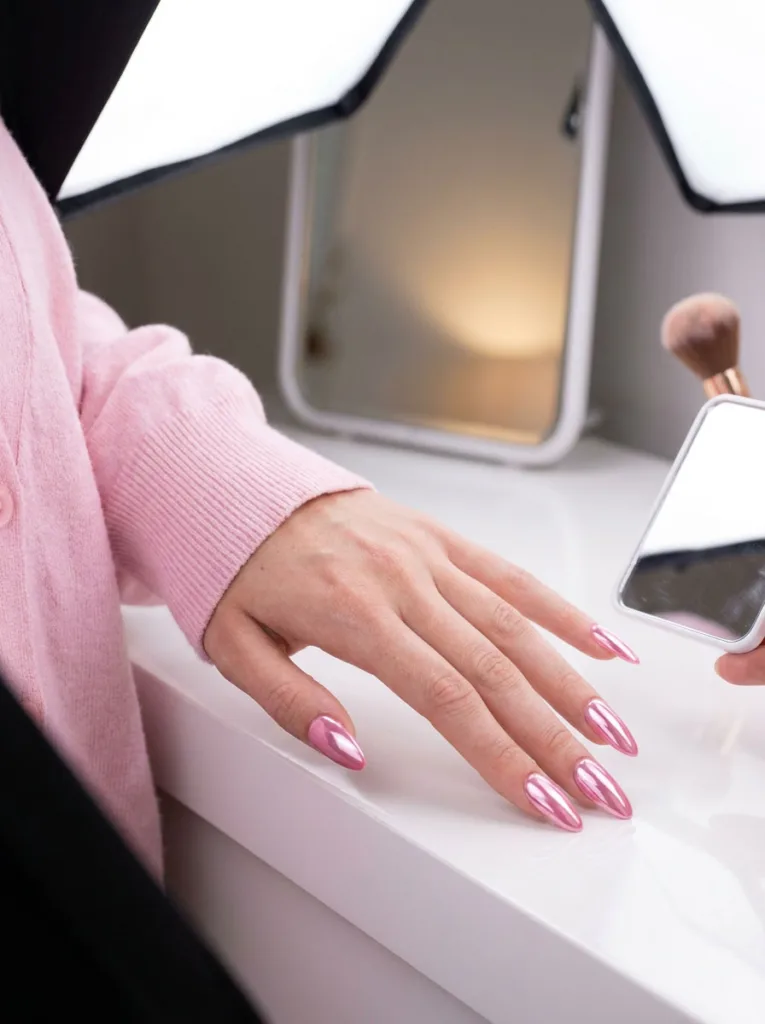 High-shine bubblegum pink chrome nails on almond-shaped nails at a vanity mirror