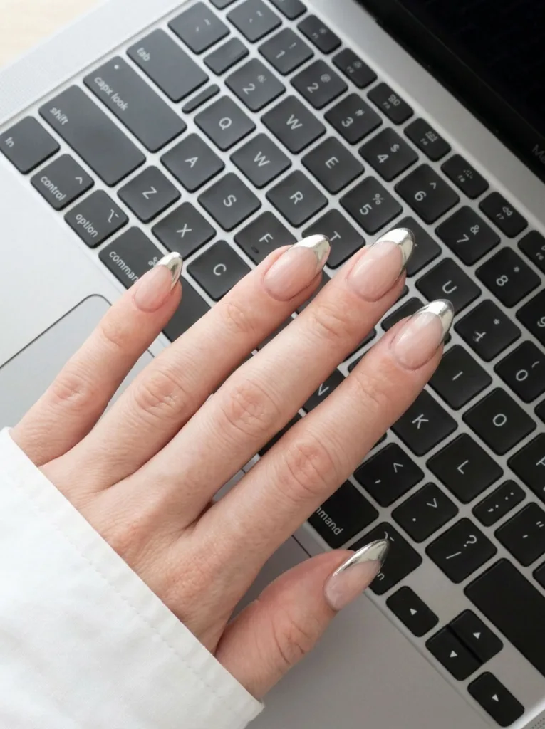 Fair-skinned hand with chrome French tip nails resting on a laptop keyboard in a bright office