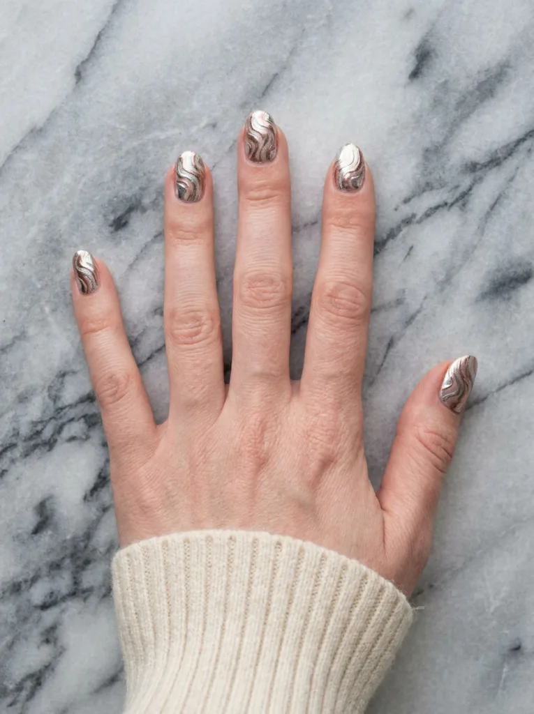 Fair-skinned hand fanned open showing silver and rose gold marbled chrome swirl nails on a marbled surface