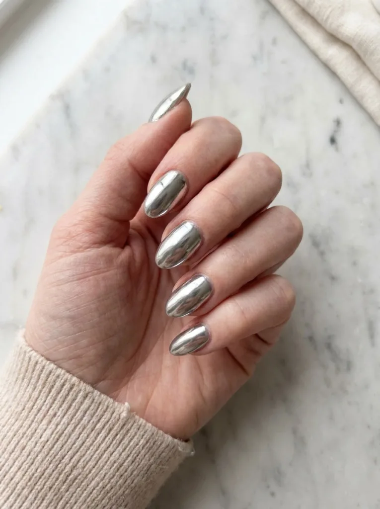 Fair-skinned hand with classic silver mirror chrome nails flat-lay on a marble countertop beside a coffee cup