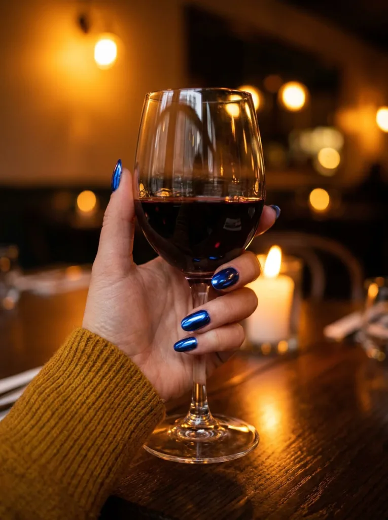 Fair-skinned hand with vivid cobalt blue chrome nails holding a glass of red wine at a restaurant table