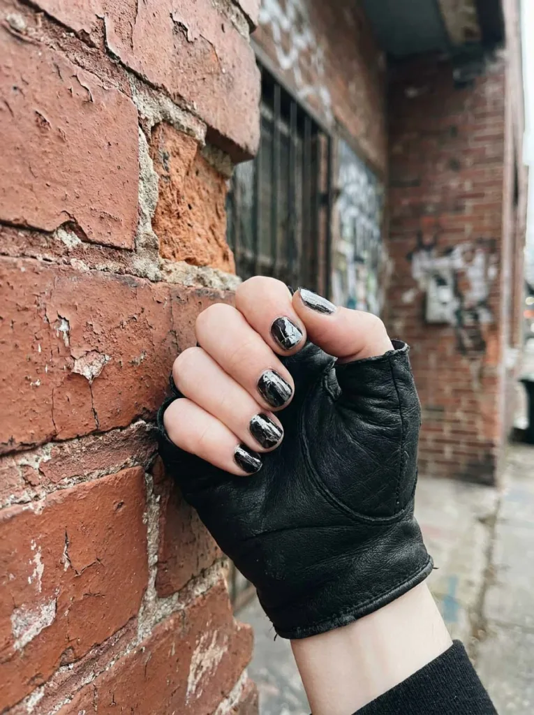 Fair-skinned hand with distressed cracked-texture black chrome nails against a weathered brick wall