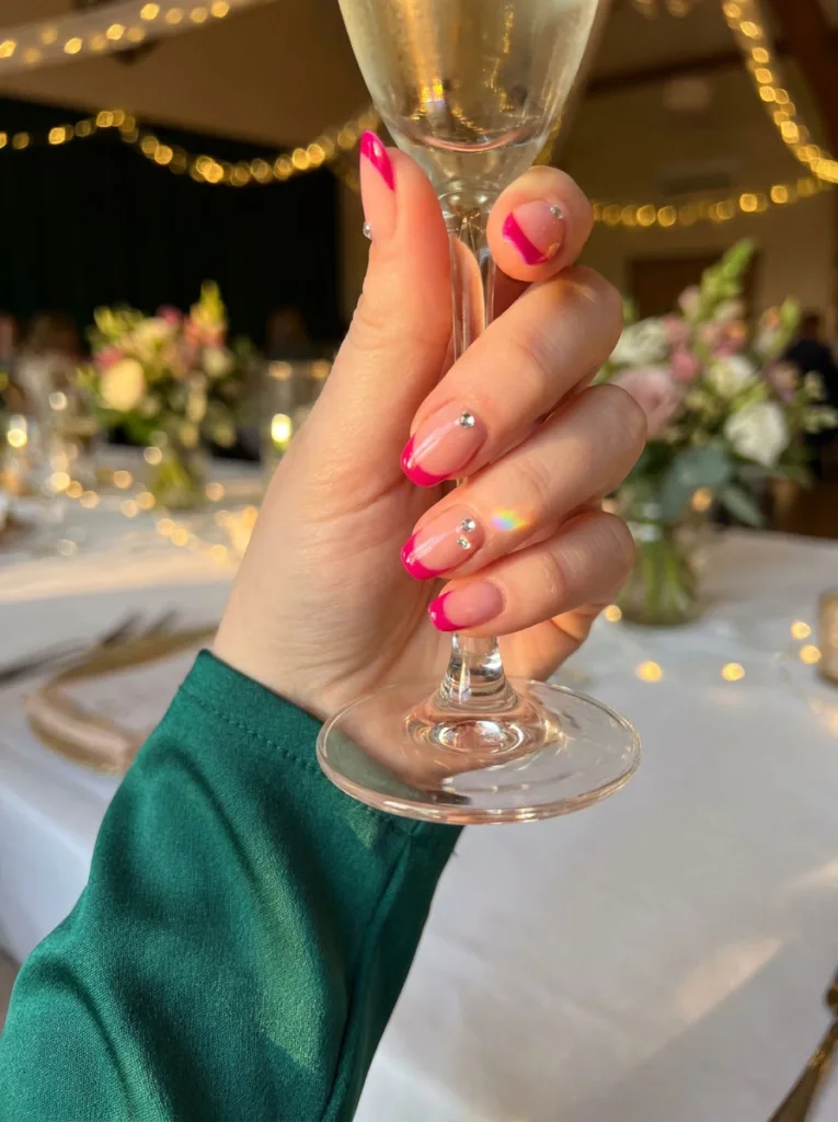 Bright fuchsia French tip nails with gem accents holding a champagne glass
