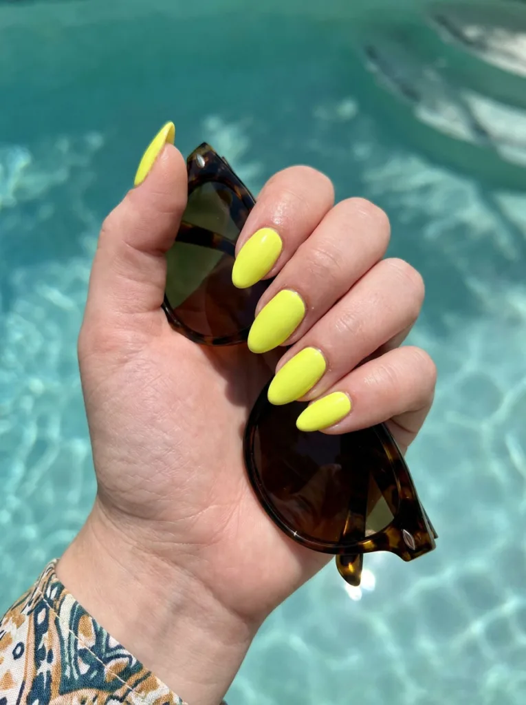 Fair-skinned hand with a bold full neon yellow manicure by a pool holding sunglasses
