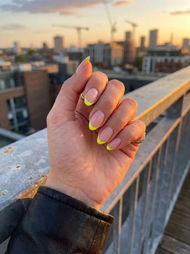 Fair-skinned hand with neon yellow micro French tip nails on a city rooftop railing