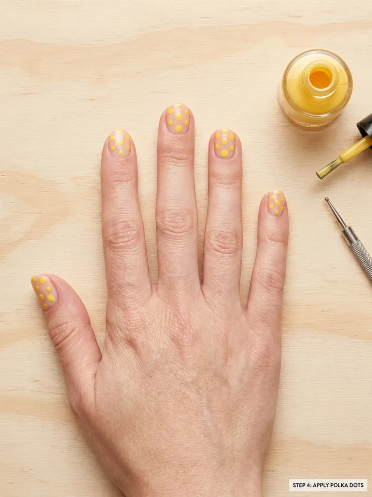 Fair-skinned hand with yellow polka dot nail art and dotting tool on a craft table
