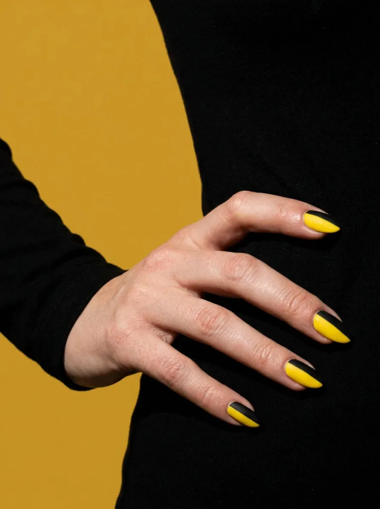 Fair-skinned hand with graphic yellow and black color block nails against a mustard backdrop