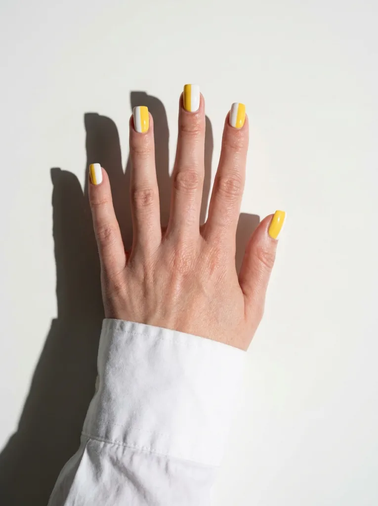 Fair-skinned hand with minimalist yellow and white geometric nails on a white desk