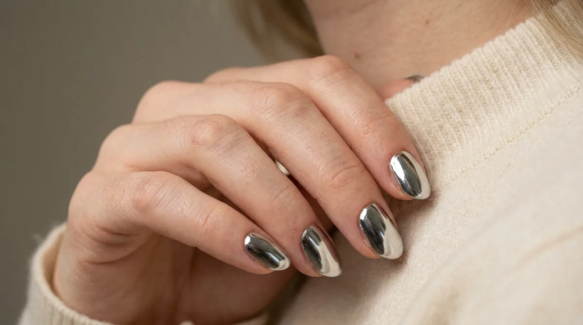 Fair-skinned hand with classic silver chrome nails, also called mirror nails, resting near the collarbone in soft studio light